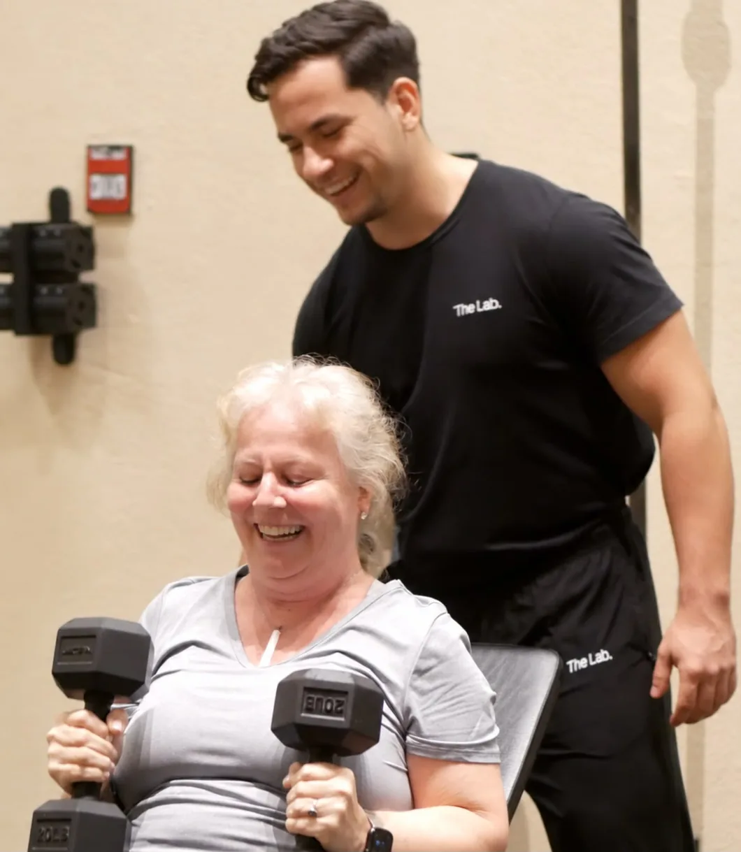 A member smiling while lifting two dumbbells, with her mentor beside her at The Lab in Hallandale Beach